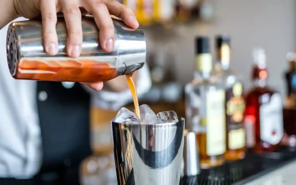 A bartender mixing whiskey and peach schnapps in a cocktail shaker filled with ice at a bar counter.